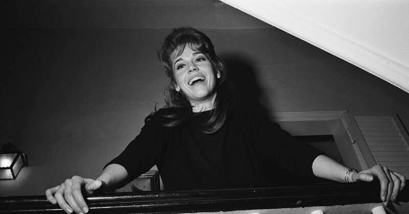 Jane Fonda laughing while leaning at the banister of the stairs in her apartment.
